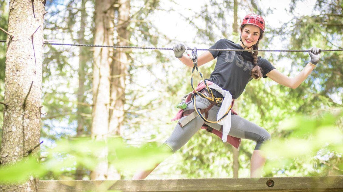 Eine abenteuerlustige Person steht auf einer Plattform zwischen Bäumen und hält sich an einem Seil fest. Sie trägt Sicherheitsausrüstung und schaut zuversichtlich in die Kamera. | © Gröbming Abenteuerpark