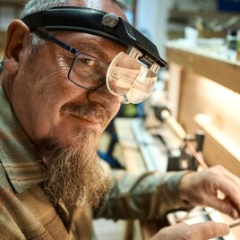 Ein älterer Mann mit einer Lupe arbeitet an einem handwerklichen Projekt. Im Hintergrund sind Werkzeuge und Materialien sichtbar.