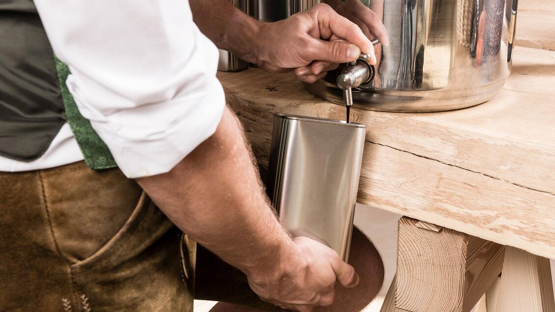 Ein Mann füllt eine Flasche aus einem Behälter. Er trägt traditionelle Kleidung und steht an einem Holztisch. | © Adamarkt Seggauberg