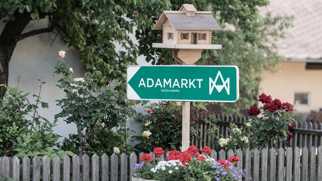 Ein Wegweiser mit der Aufschrift "ADAMARKT" steht neben einem kleinen Vogelhaus. Blumen und eine Holzlattenzaun umgeben die Szene. | © Adamarkt Seggauberg