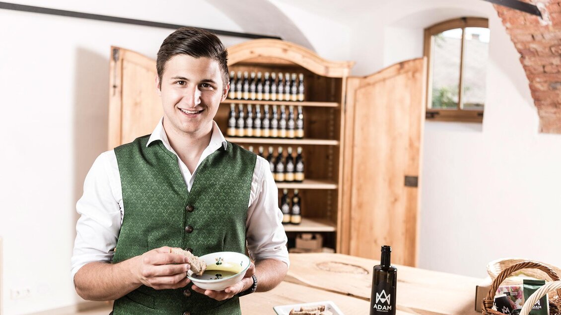 Ein junger Mann in traditioneller Kleidung hält eine Schüssel mit Essen in der Hand. Im Hintergrund sind Regale mit Flaschen und ein Holzschrank zu sehen. | © Adamarkt Seggauberg