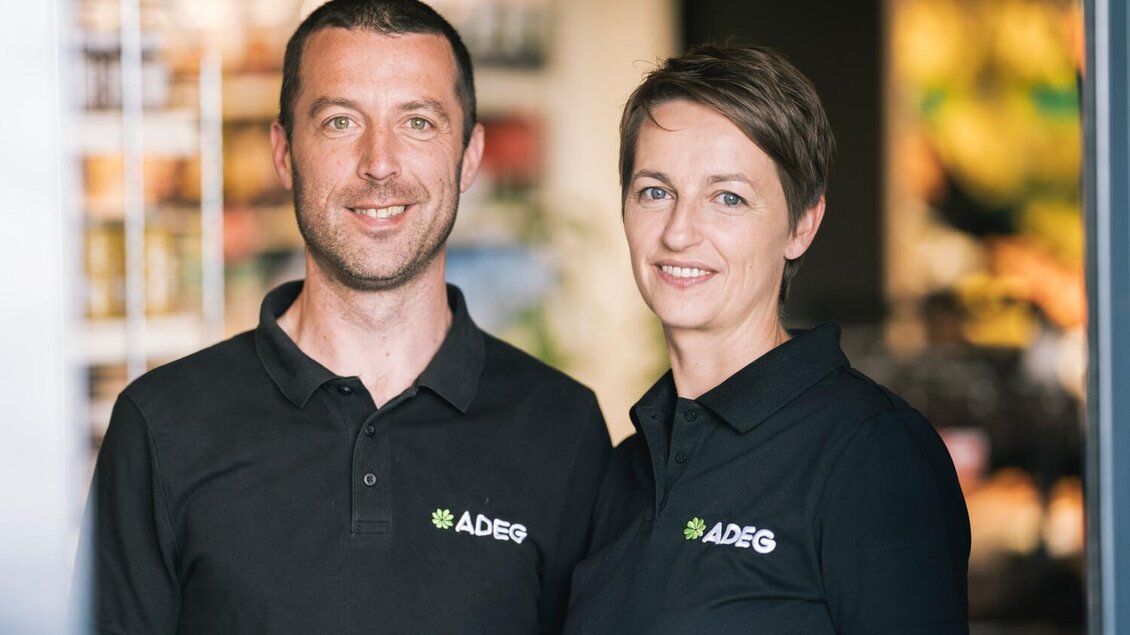 Ein freundliches Paar steht in einem Verkaufsraum. Beide tragen schwarze Poloshirts mit dem Logo von ADEG. | © Stefan Leitner