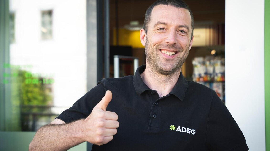 Ein fröhlicher Mann mit Daumen hoch steht vor einem Geschäft. Er trägt ein schwarzes Poloshirt mit dem Logo von ADEG. | © Stefan Leitner
