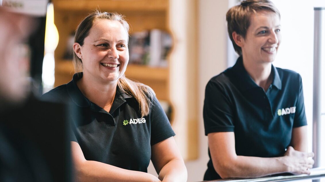 Zwei lächelnde Frauen stehen an einem Empfangstresen. Beide tragen schwarze Poloshirts mit dem Logo von ADEG. | © Stefan Leitner