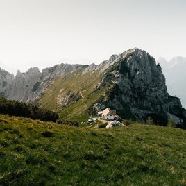 An impressive mountain landscape with a steep rock formation. In the foreground, there is a small house amidst green meadows.