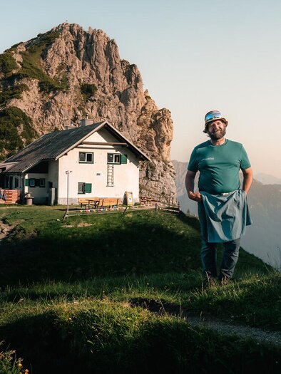 Ein Mann in blauer Kleidung steht vor einem typischen Berghaus. Im Hintergrund sind majestätische Berge zu sehen und die Landschaft ist grün und hügelig.