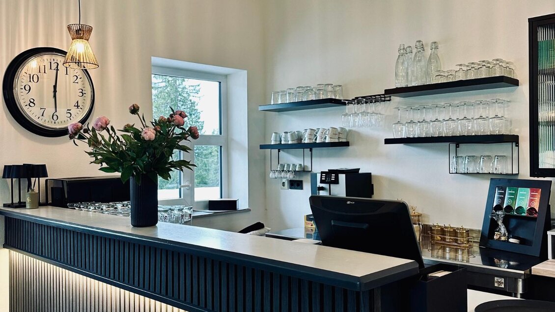 Ein modernes Café mit einem eleganten Tresen und einer schönen Blume in einer Vase. Im Hintergrund sind Gläser und eine große Uhr an der Wand zu sehen. | © Gipfelmomente