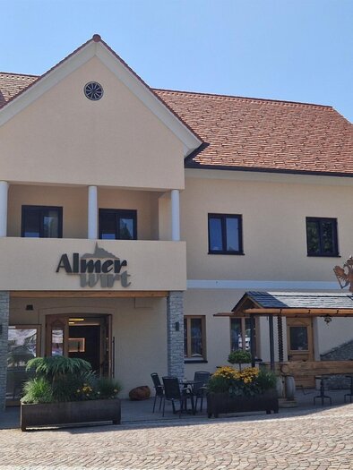 A pretty building with a restaurant and an outdoor terrace. In the background, there is a church tower surrounded by trees. | © Almer