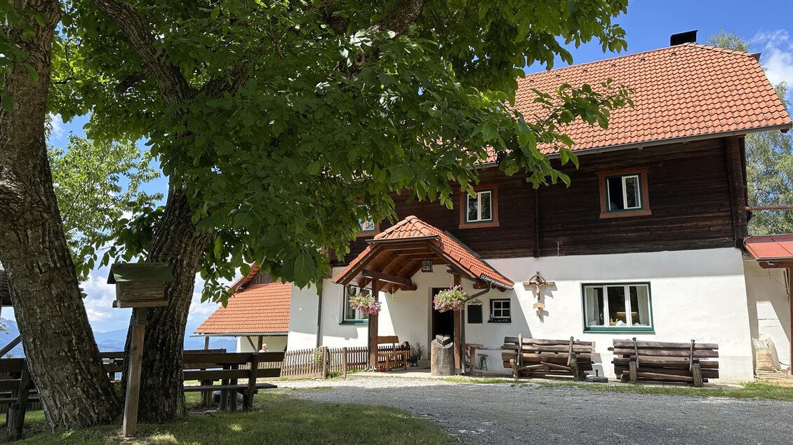 Ein gemütliches Holzhaus mit rotem Dach, umgeben von Bäumen. Der Eingang ist einladend und es gibt Sitzgelegenheiten im Freien. | © TV Hochsteiermark