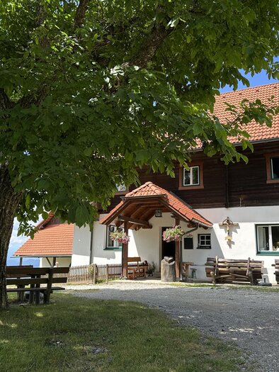 A cozy wooden house with a red roof, surrounded by trees. The entrance is inviting and there are outdoor seating areas. | © TV Hochsteiermark