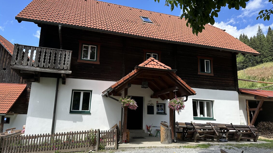 Ein rustikales Holzhaus mit rotem Dach und weißen Wänden. Vor dem Haus steht eine kleine Terrasse mit Bänken und einem schönen Ausblick ins Grüne. | © TV Hochsteiermark