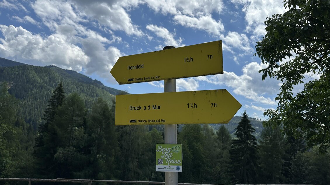 Ein Wegweiser im Freien zeigt Richtung und Entfernung zu verschiedenen Zielen. Im Hintergrund sind Bäume und Berge zu sehen, bei klarem Himmel. | © TV Hochsteiermark