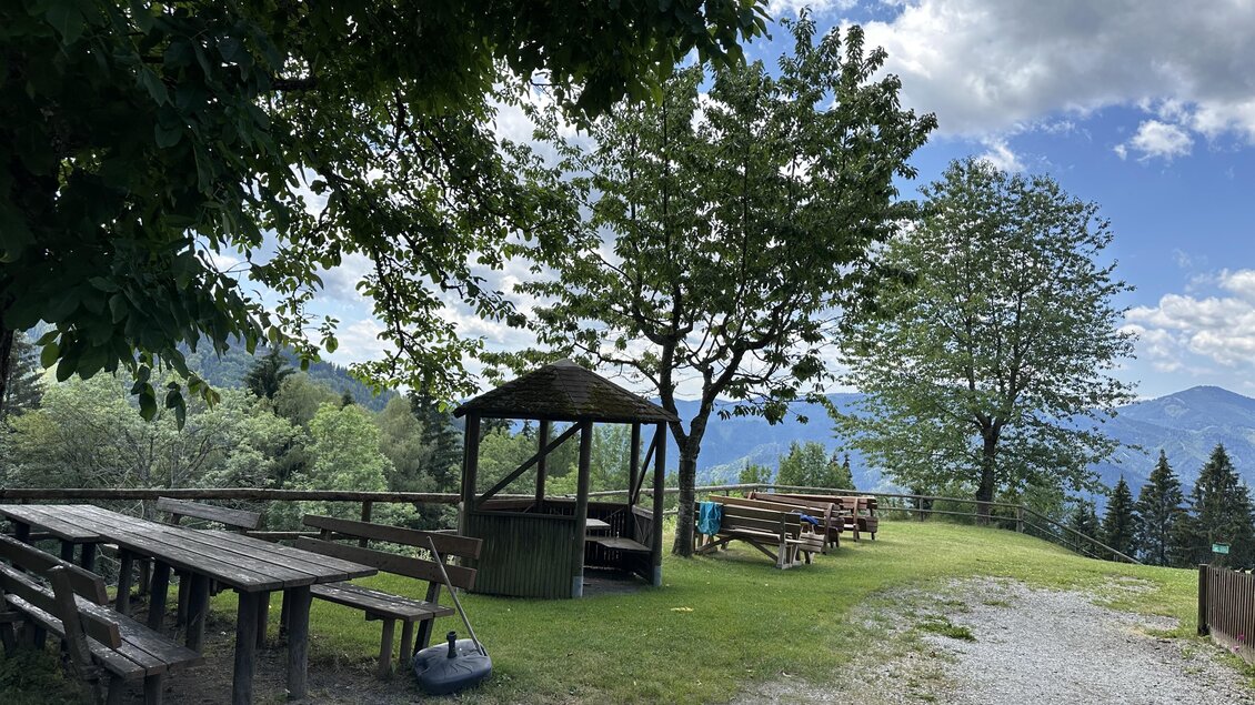 Eine malerische Aussicht mit Holztischen und Bänken unter Bäumen. Der Himmel ist teilweise bewölkt und die Landschaft ist grün. | © TV Hochsteiermark