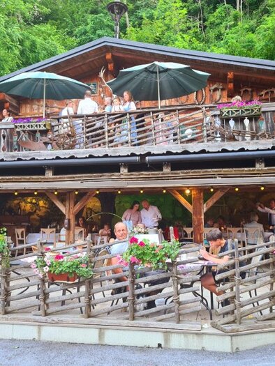 Ein gemütliches Restaurant mit Holzveranda inmitten der Natur. Viele Gäste genießen das Essen bei schönem Wetter und blühenden Pflanzen. | © Almhütte