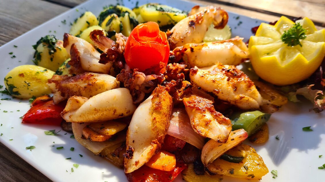 Ein schön angerichteter Teller mit gegrilltem Oktopus, buntem Gemüse und Kürbisscheiben. Dazu gibt es eine Zitrone und eine rote Blume als Dekoration. | © Almhütte