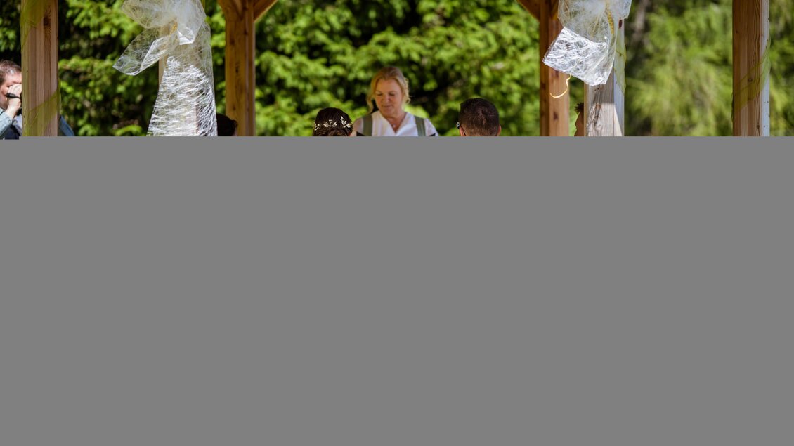 Eine Hochzeitszeremonie unter einem Holzpavillon. Das Brautpaar sitzt auf liebevoll dekorierten Stühlen, während ein Zeremonienleiter vor ihnen spricht. | © Almidylle Sabathy