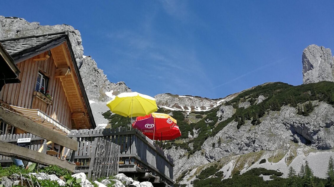 Eine Berghütte mit bunten Sonnenschirmen steht vor einer beeindruckenden Felsenlandschaft. Der Himmel ist klar und die Berglandschaft wirkt einladend. | © Veronika Grill