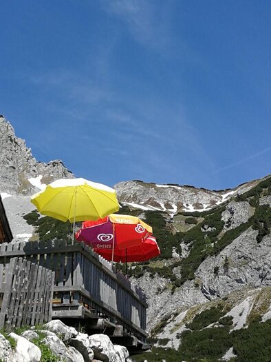 Eine Berghütte mit bunten Sonnenschirmen steht vor einer beeindruckenden Felsenlandschaft. Der Himmel ist klar und die Berglandschaft wirkt einladend. | © Veronika Grill