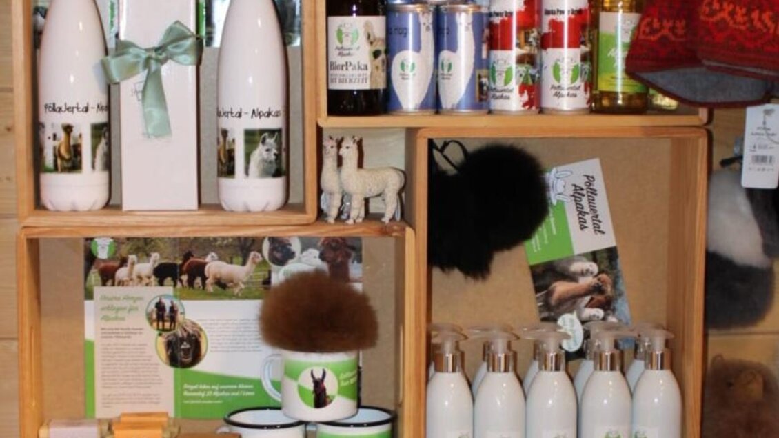 Ein Regal mit verschiedenen alpaka-inspirierten Produkten, darunter Trinkflaschen, Tassen und Geschenksets. Die Artikel sind ordentlich in Holzkisten arrangiert. | © Ganster