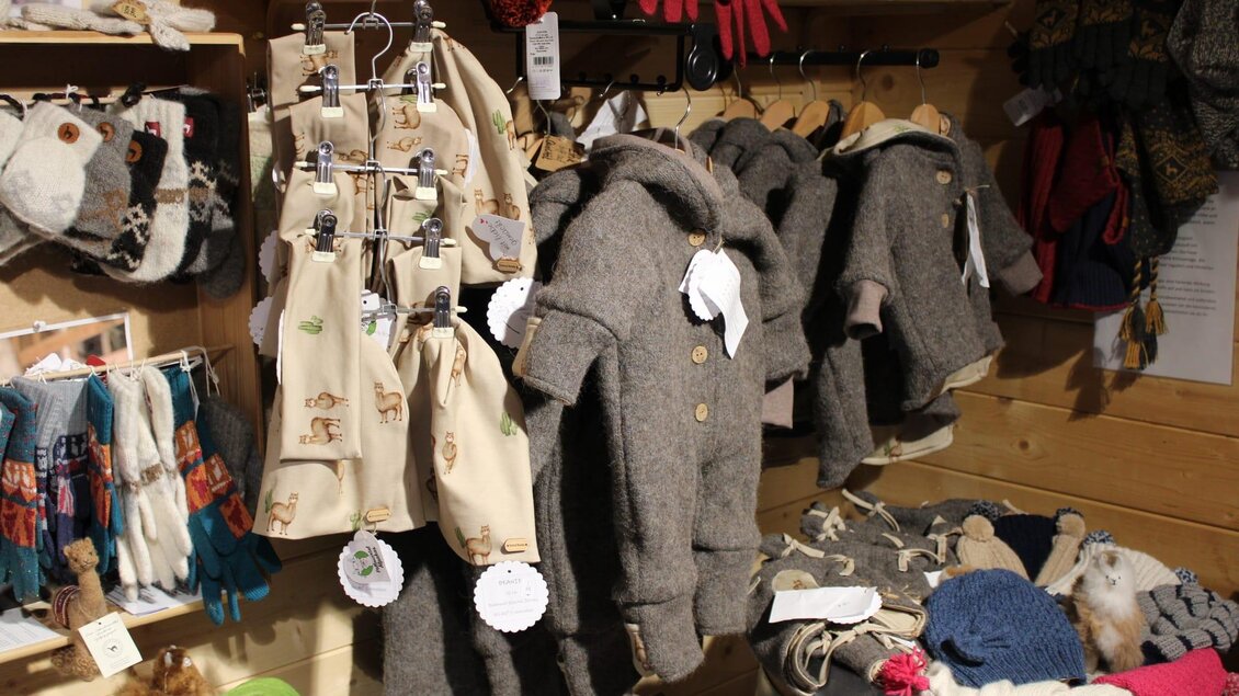 Eine Auswahl an Kleidung für Kinder, darunter graue Mäntel und verschiedene Accessoires. Die Sachen hängen an einer Holzwand in einem gemütlichen Laden. | © Ganster