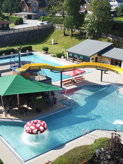 Ein großer Outdoor-Wasserpark mit einem Schwimmbecken, einer Rutsche und einer Lounge. Rund um den Pool gibt es grüne Wiesen und gemütliche Liegestühle. | © Rainer & Steiner GmbH