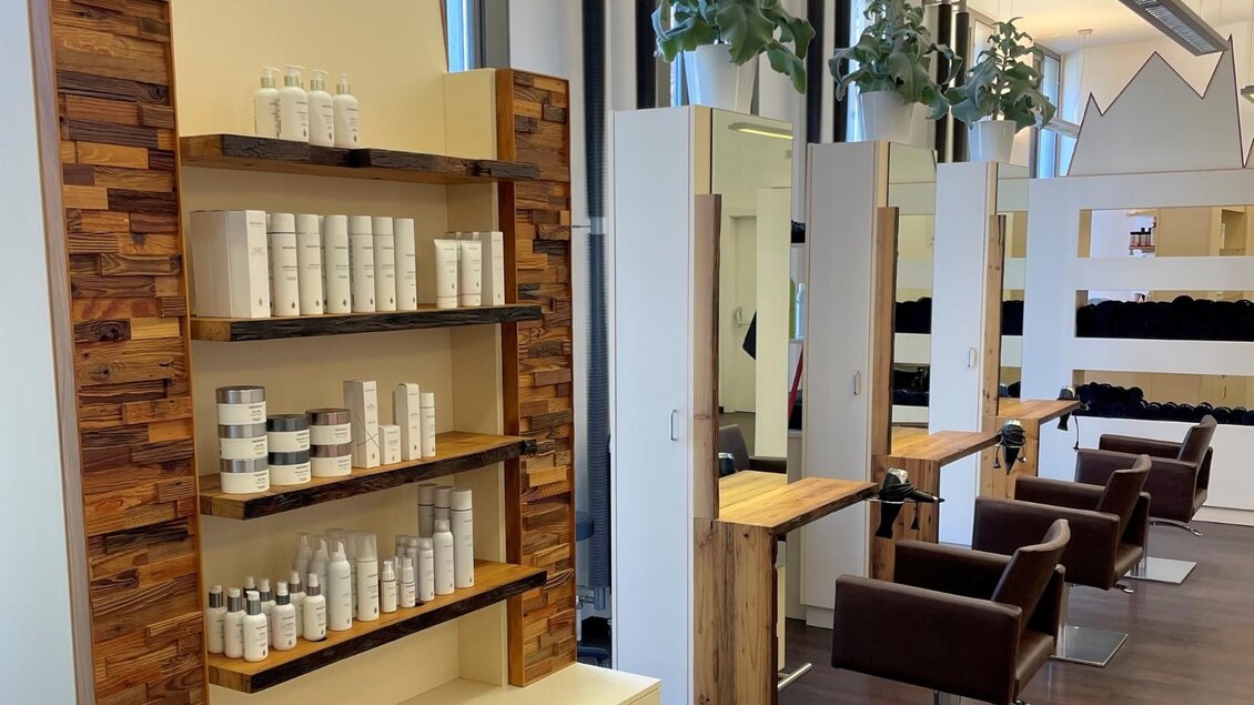 Ein moderner Friseursalon mit einem Regal für Pflegeprodukte und Friseurstühlen. Große Fenster sorgen für viel Tageslicht. | © Alpenhaar Bad Aussee