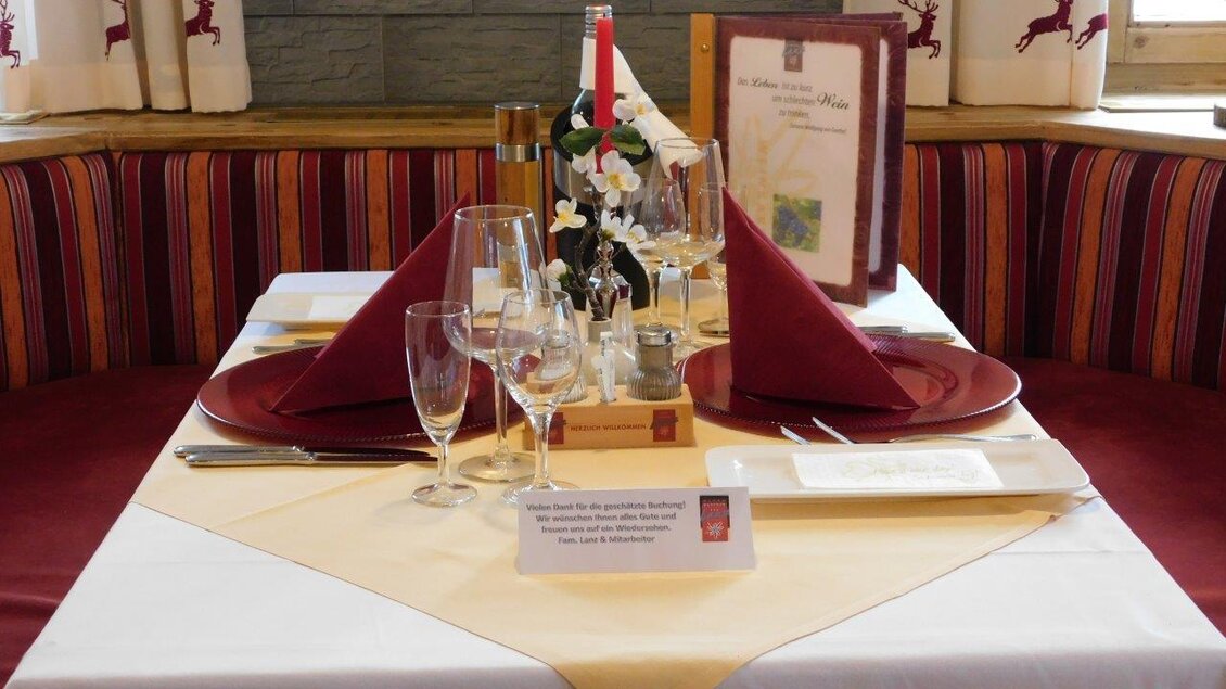 Ein schön gedeckter Tisch in einem Restaurant mit roten Servietten und Gläsern. Die Tischdeko umfasst eine kleine Blume und eine Kerze. | © Alpenhotel Lanz
