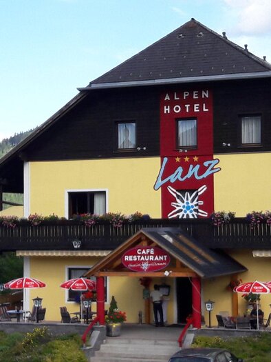 A charming alpine hotel with colorful flower windows. In front of the hotel, there is an inviting café with umbrellas and a beautiful view of the mountains. | © Alpenhotel Lanz