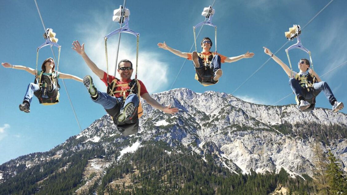 Vier Personen fahren fröhlich an einer Seilbahn über eine majestätische Berglandschaft. Die hohe Bergkulisse und der klare Himmel schaffen eine aufregende Atmosphäre. | © Alpincenter Dachstein