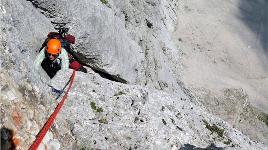Ein kletternder Bergsteiger mit einem orangefarbenen Helm schwingt sich an einem Seil über graue Felsen. Im Hintergrund ist eine weite, meist leere Landschaft zu sehen. | © Alpincenter Dachstein