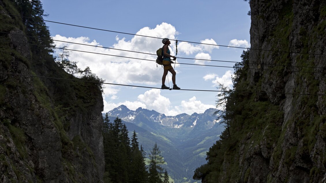 Eine Person balanciert auf einem Drahtseil zwischen zwei Felsen. Im Hintergrund sind Berge und ein blauer Himmel mit Wolken zu sehen. | © Alpincenter Dachstein