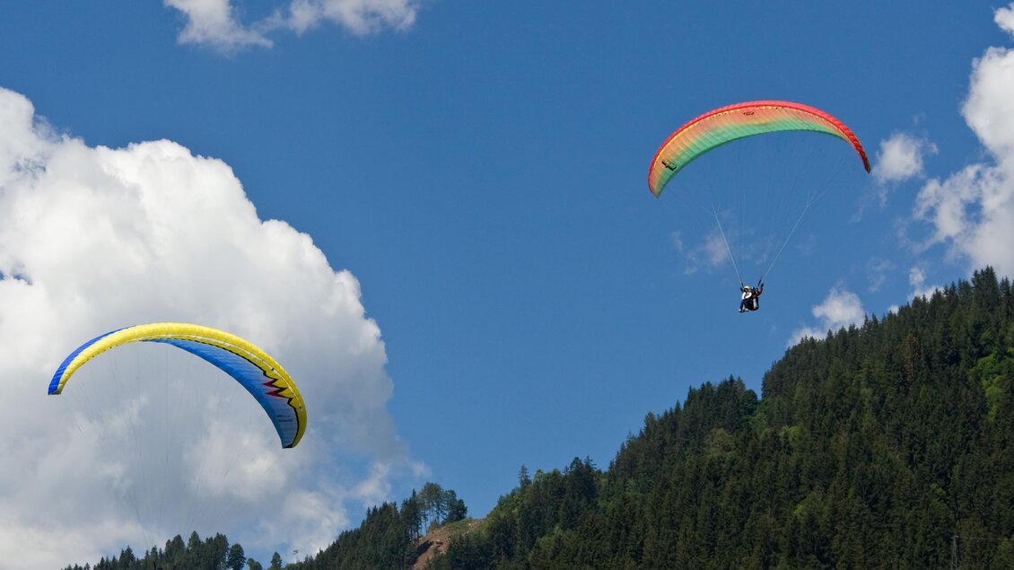Zwei Gleitschirmflieger schweben über einem grünen Berg mit strahlend blauem Himmel. Weiße Wolken ergänzen die malerische Landschaft. | © Alpincenter Dachstein