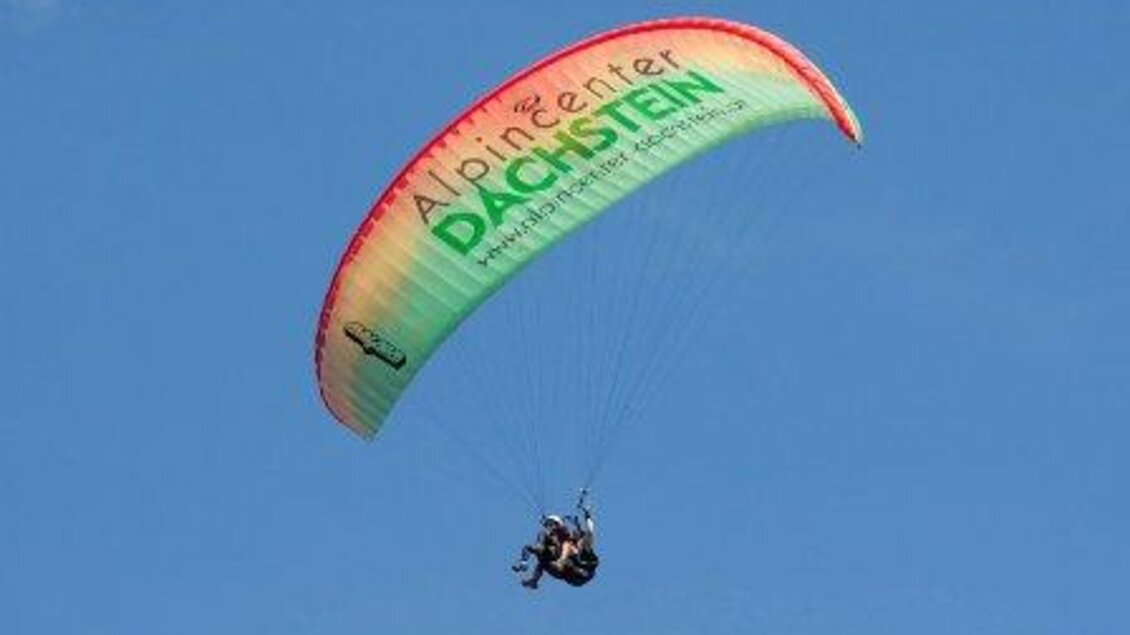 Ein Fallschirmspringer schwebt mit einem bunten Gleitschirm am blauen Himmel. Der Schirm hat die Aufschrift "Alpincenter Dachstein". | © Alpincenter Dachstein