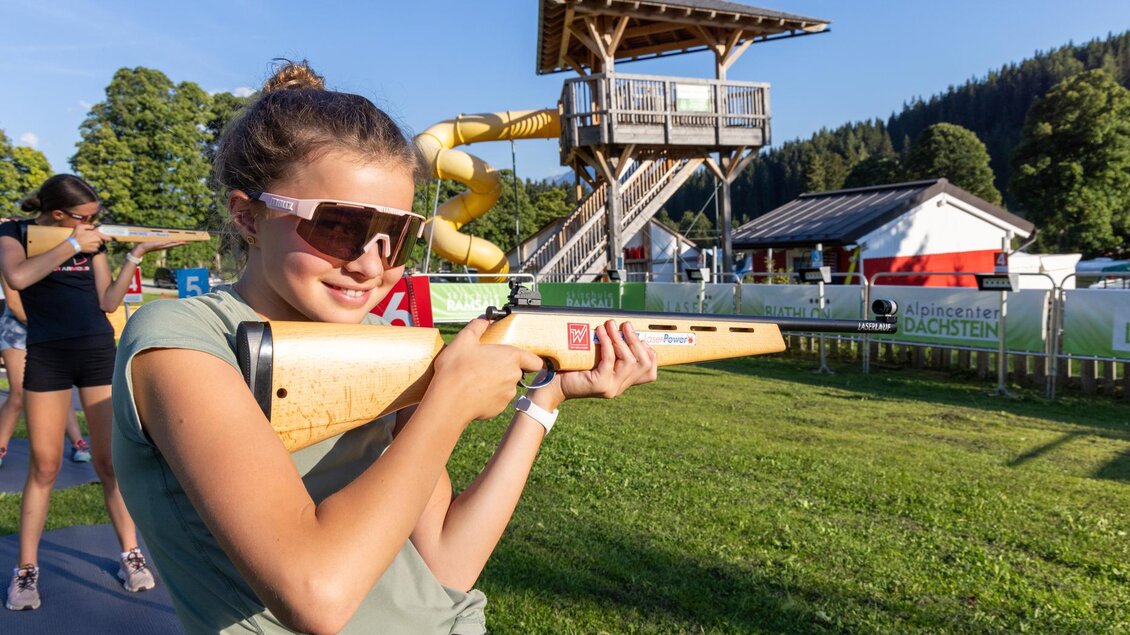 Ein junges Mädchen zielt mit einem Sportgewehr auf eine Zielscheibe. Im Hintergrund ist ein Spielturm mit einer Rutsche zu sehen. | © Alpincenter Dachstein