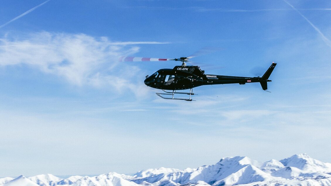 Ein Hubschrauber fliegt über schneebedeckte Berge. Der Himmel ist blau mit einigen Wolken. | © Alpincenter Dachstein