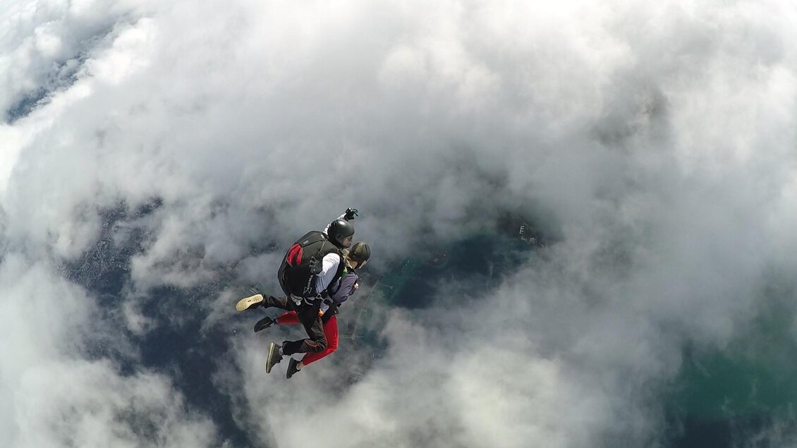 Ein Sprung aus  der Höhe mit zwei Fallschirmspringern inmitten von Wolken. Die Landschaft darunter ist teilweise sichtbar und bietet eine beeindruckende Aussicht. | © Alpincenter Dachstein