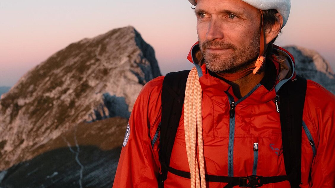 Ein Bergsteiger steht in der Berglandschaft mit einem Kletterhelm und einer roten Jacke. Der Sonnenuntergang färbt den Himmel in sanften Tönen. | © TV Gesäuse