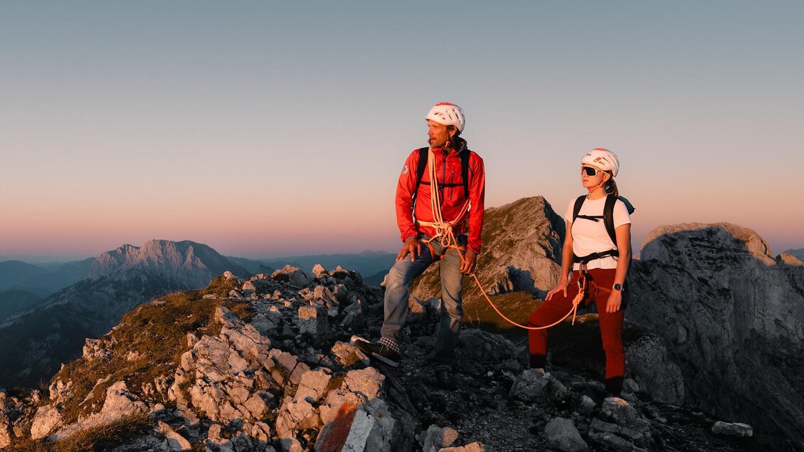 Ein Kletterer und eine Kletterin stehen auf einem felsigen Gipfel. Im Hintergrund sind sanfte Berge und ein farbenfroher Himmel zu sehen. | © TV Gesäuse