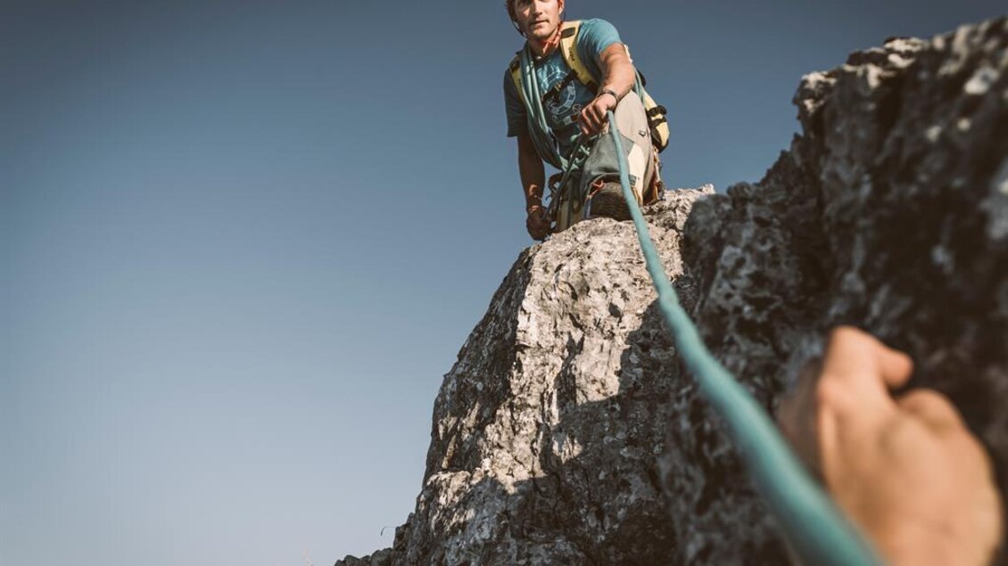 Ein Kletterer steht oben auf einem Felsen und sichert seinen Partner. Die Umgebung zeigt hohe Berge und einen klaren Himmel. | © Stefan Leitner