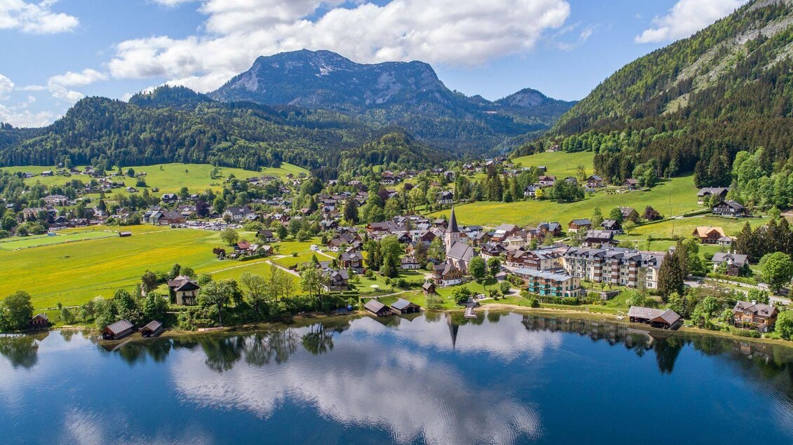 <p>Die malerische Landschaft in Altaussee mit dem klaren Altausseer See, umgeben von grünen Wiesen und den Bergen des Ausseerlandes. Im Hintergrund ist der Ort mit seinen traditionellen Gebäuden zu sehen.</p> | © Drohnenfotos.at-Andreas Tischler