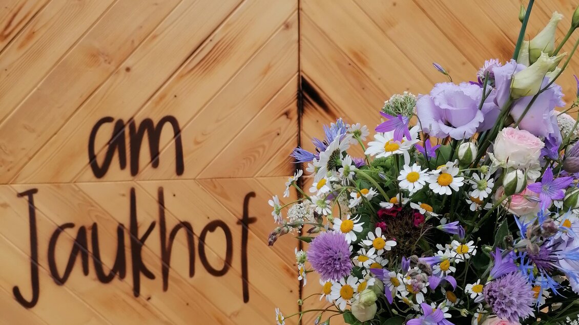 Ein bunter Blumenstrauß wird vor einer Holzwand mit der Aufschrift "am Jaukohf" gehalten. Die Wand gehört zur Naturwerkstatt. | © Familie Stiendl