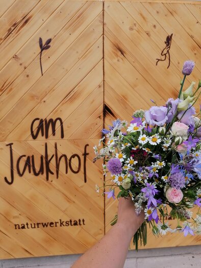 A colorful bouquet is held in front of a wooden wall with the inscription "am Jaukohf." The wall belongs to the nature workshop. | © Familie Stiendl