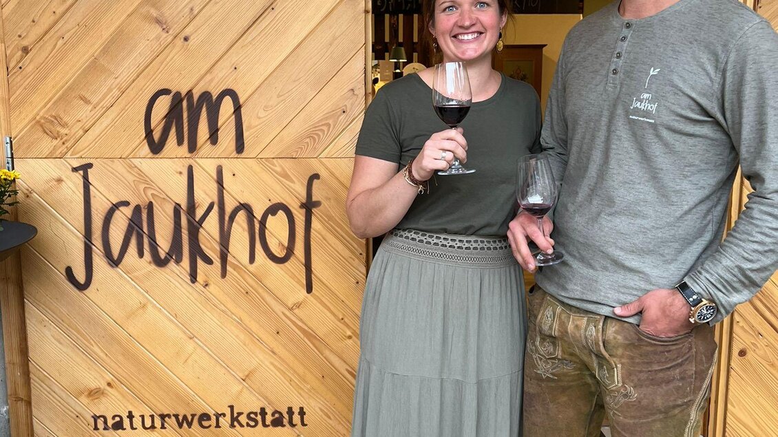 Ein Paar steht vor einer Holztür mit dem Schriftzug "am Jaukhof". Sie halten Gläser in der Hand und lächeln freundlich. | © Familie Stiendl