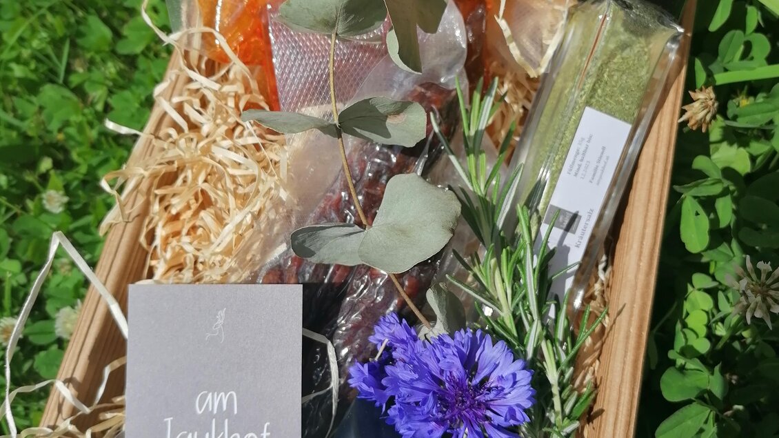 Ein dekorierter Geschenkkarton mit verschiedenen Produkten, darunter ein Glas mit einer Kerze und eine Flasche. Eingerahmt von buntem Füllmaterial und frischen Kräutern. | © Familie Stiendl