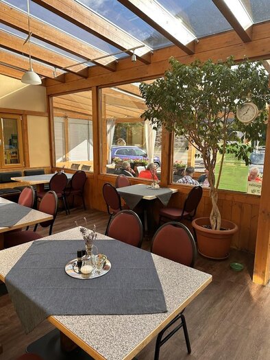 Ein gemütliches Restaurant mit Holzmöbeln und großen Fenstern. Pflanzen und gedeckte Tische schaffen eine einladende Atmosphäre.