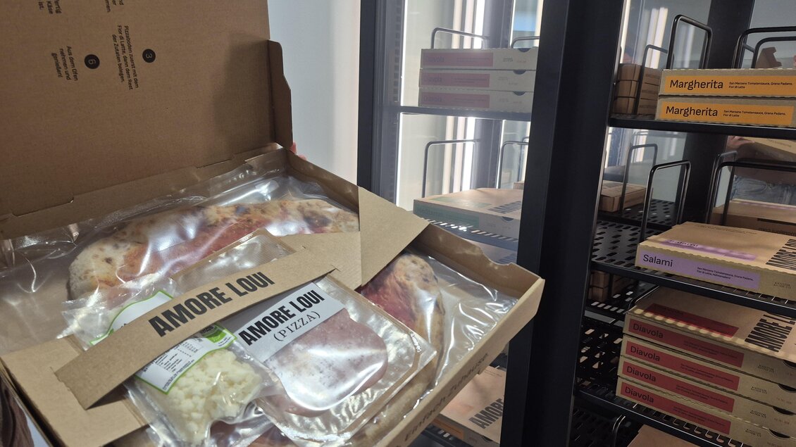 Eine geöffneten Pizzakarton mit verpackter Pizza. Im Hintergrund sind weitere Pizzakartons in einem Kühlschrank zu sehen. | © TV Hochsteiermark