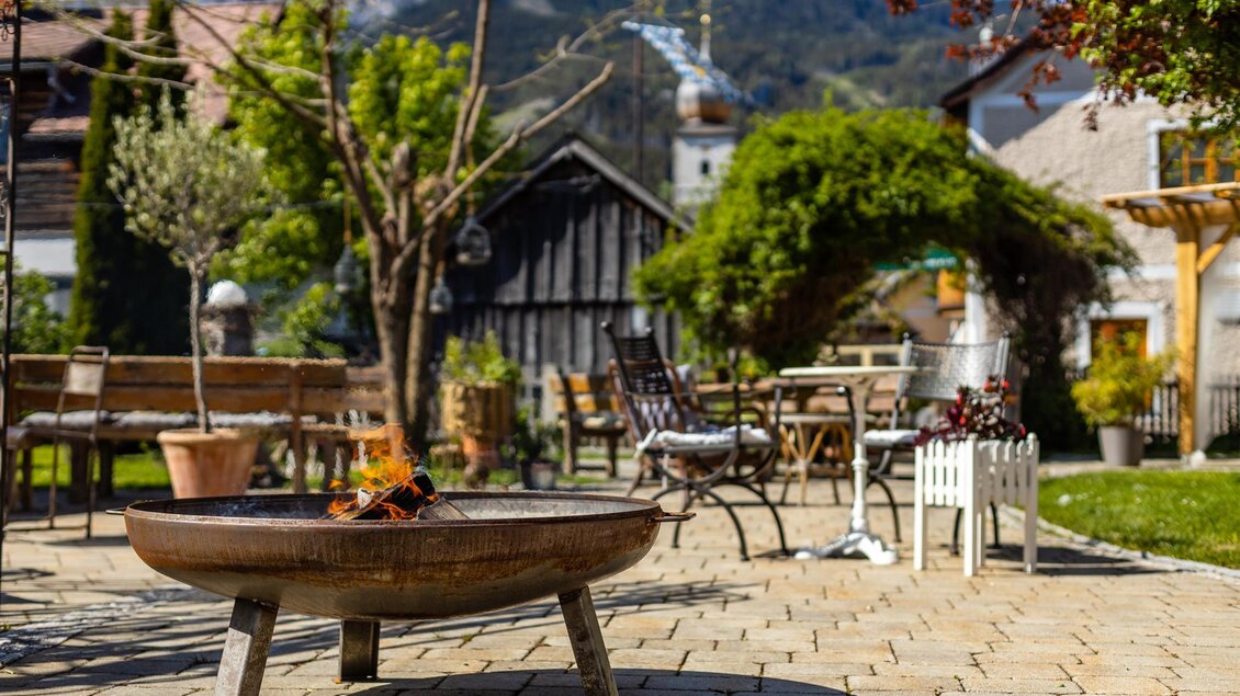 Ein schöner Garten mit einem Feuerring im Vordergrund. Im Hintergrund sind Sitzgelegenheiten und Bäume sichtbar, umgeben von einer malerischen Landschaft. | © Gustav Neubauer