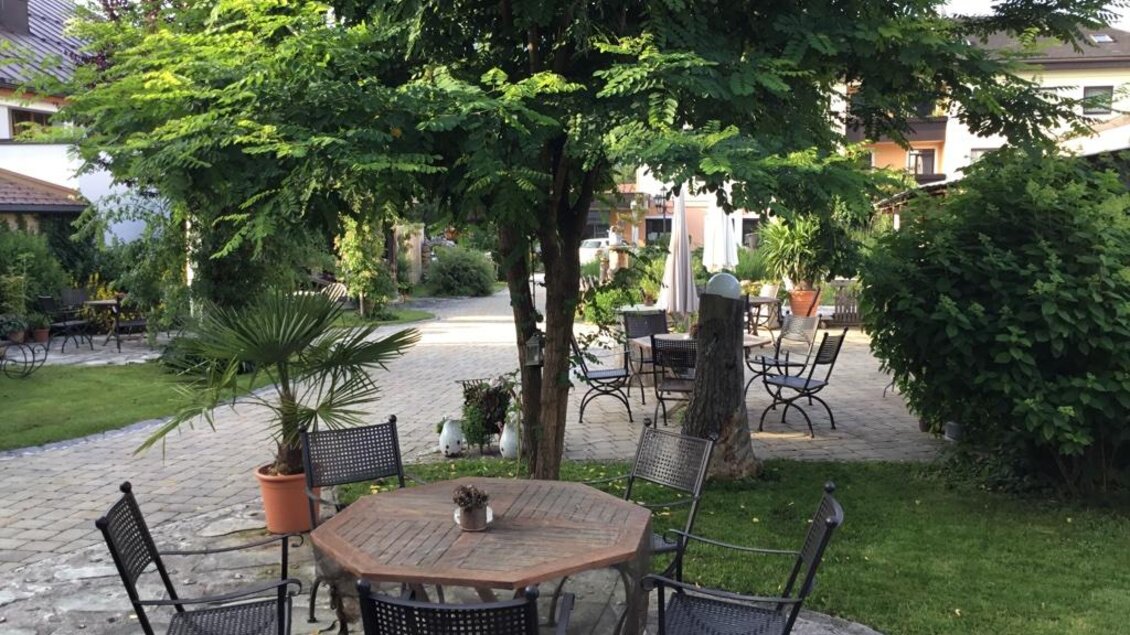 Ein einladender Garten mit einem runden Tisch und Stühlen unter einem grünen Baum. Im Hintergrund sind weitere Sitzgelegenheiten und fruchtbares Grün zu sehen. | © Gustav Neubauer