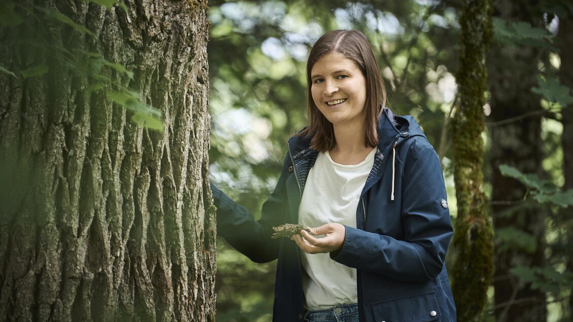 Eine Frau steht neben einem Baum im Wald und lächelt. Sie hält etwas in der Hand und trägt eine Regenjacke. | © Nationalpark Gesäuse