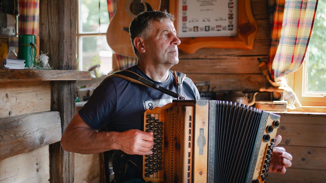 Ein Mann spielt Akkordeon in einer gemütlichen, holzverkleideten Stube. Im Hintergrund sind Fenster mit bunten Vorhängen und eine Gitarre zu sehen.
